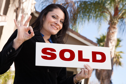 Happy Attractive Hispanic Woman Holding Sold Sign and In Front of House.